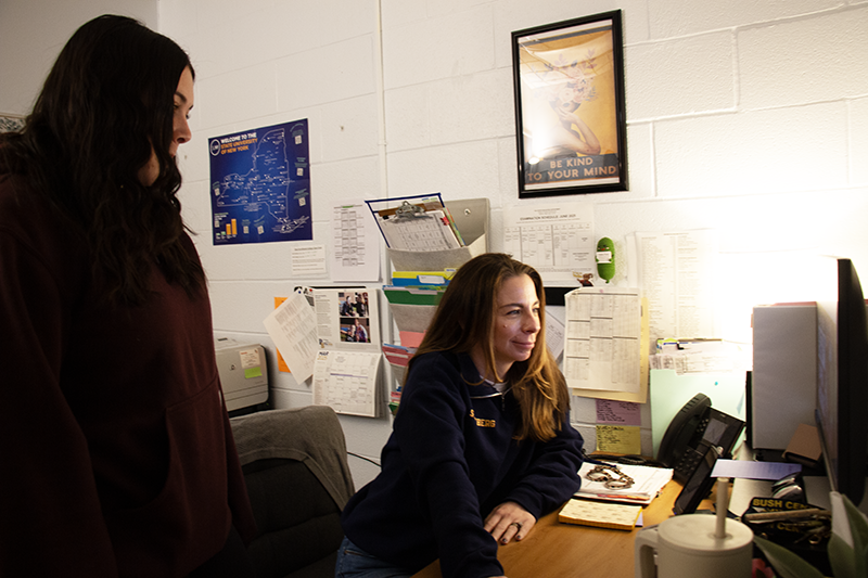 A woman sits at a desk looking at a computer. A high school student stands next to her watching.