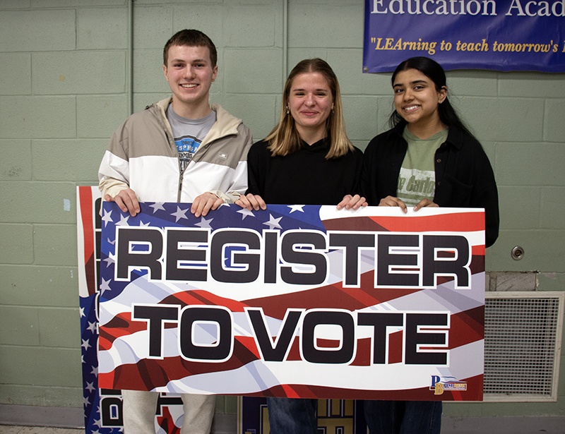 Three high school kids - a boy and two girls, from left to right, smile and hld up a large sign that says Register to Vote with an American flag background.