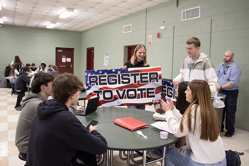 Two high school kids stand near a round table where other high school kids are sitting. The standing kids are holding a large sign that says Register to Vote with a US flag as the background.