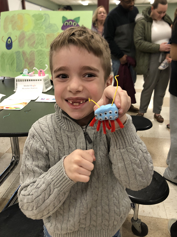 A little boy smiles as he holds up an art project he made.