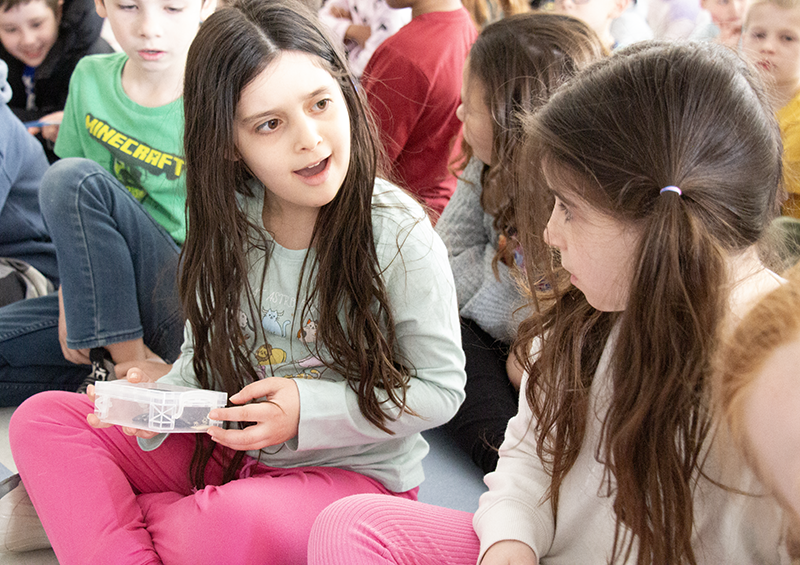 Two little girls sitting on the floor hold a plastic container that has a dinosaur artifact in it. They are looking at each other and smiling.