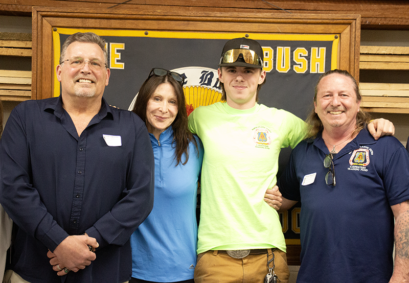 Two men on the ends and a woman wearing a blue shirt stnad next to a young man wearing a green tshirt and black ball cap. They are all smiling.