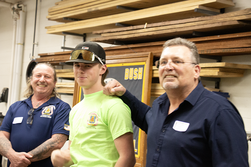 Two men stand next to a high school boy, wearing a green tshirt and a black baseball cap. One man has his hand on the boy's shoulder. They are smiling.