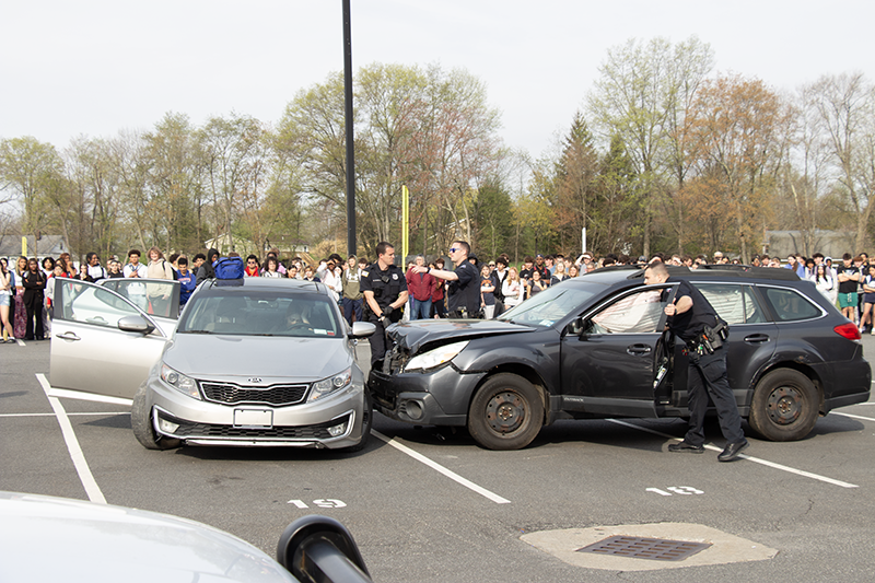 Two wrecked cars sit in a parking lot with lots of people around them. It is a mock accident.