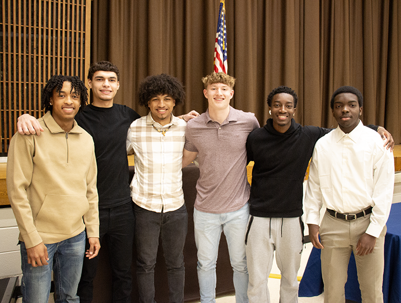 Six high school boys, all dressed nicely, stand together arm in arm, in an auditorium.