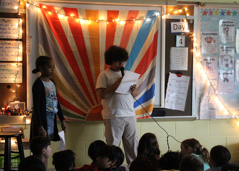 A fourth-grade boy reads a poem that he wrote.  Behind him is a multi-colored striped background. A fourth-grad girl is stand just to the left of him.