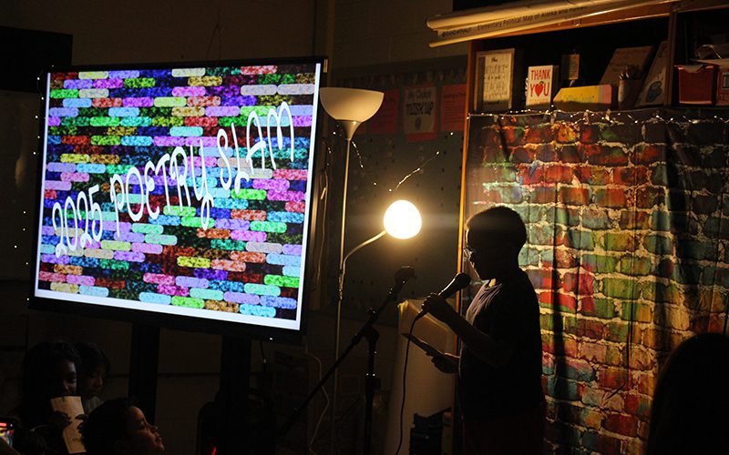 In a darkened room with a spotlight on him, a fourth-grade boy reads a poem at a microphone. Next to him is a multicolored lit sign that says Poetry Slam.