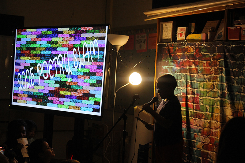 In a darkened room with a spotlight on him, a fourth-grade boy reads a poem at a microphone. Next to him is a multicolored lit sign that says Poetry Slam.