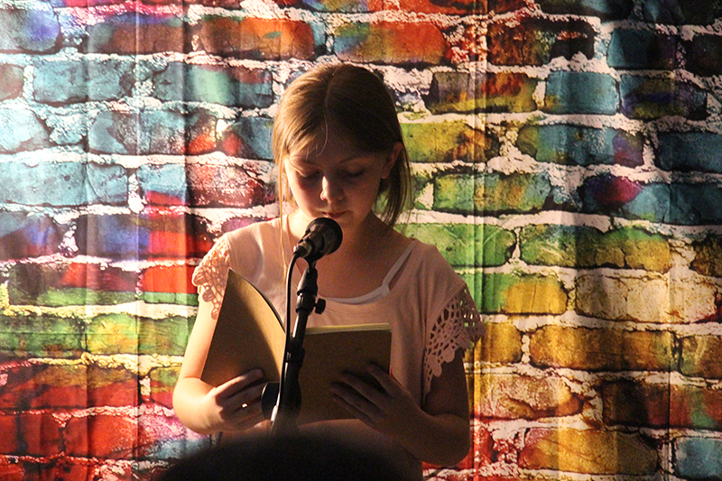 With a fabric, multi-colored brick background, a fourth-grade girl stands at a microphone and recites a poem that she wrote. It is a darkened room.