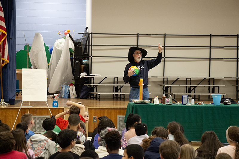 a man wearing black and a black wizard hat has one hand up and holds a ball in the other.