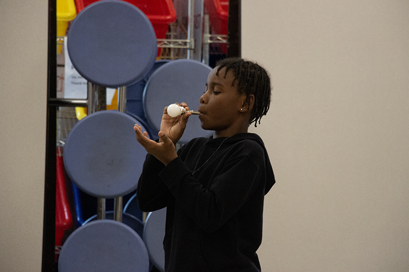 An elementary kid holds a trumpet mouthpiece and blows into it, keeping a ball balanced on it.