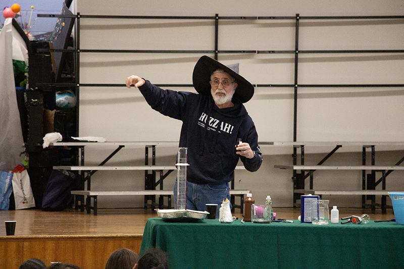 a man wearing black and a black wizard hat talks to kids from behind a table with science equipment on it.