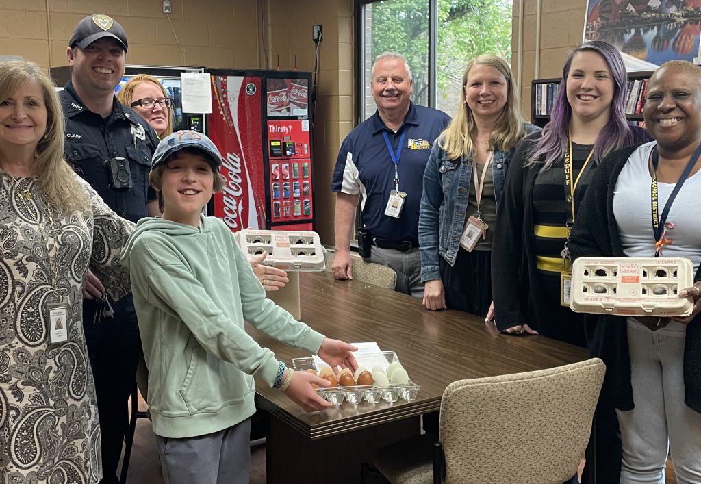 A group of seven adults stand around atable with cartons of eggs. A middle school boy stands in front, waring a light hoodie and a cap smiling. He is also holding eggs.