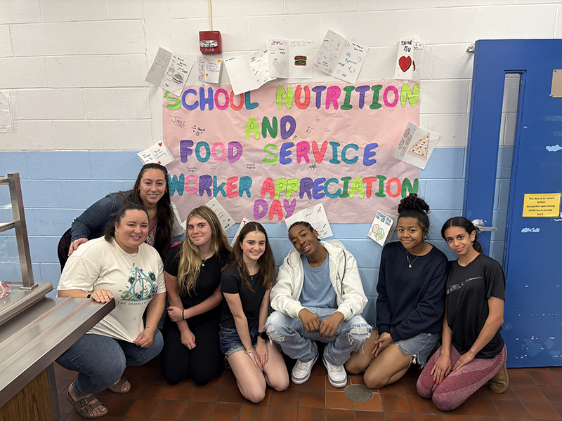 A colorful banner hangs on the wall that says School Nutrition and Food Service Worker Appreciation day. In front are seven students and staff members.