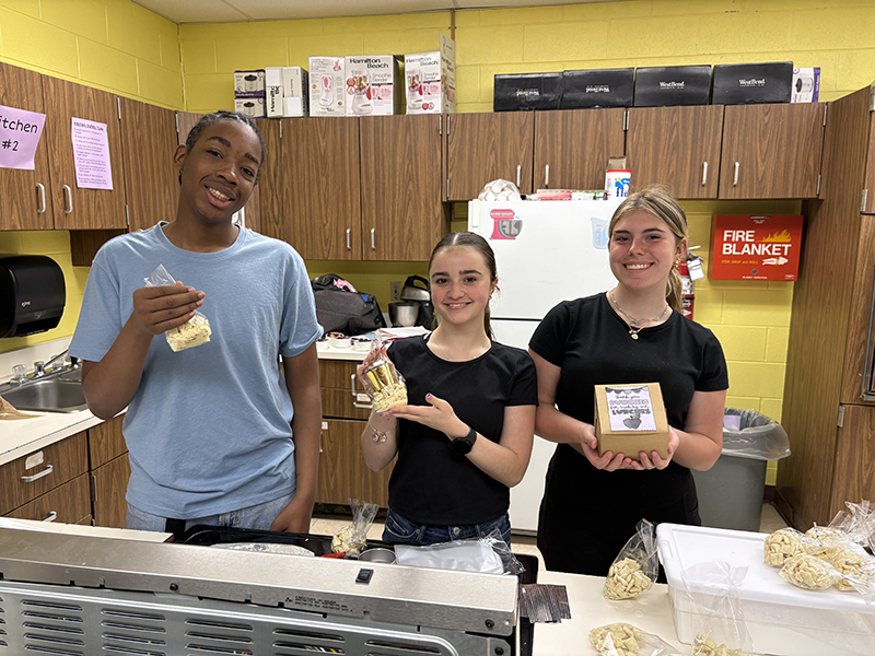 Three middle school students stand in a kitchen holding u treats they made.
