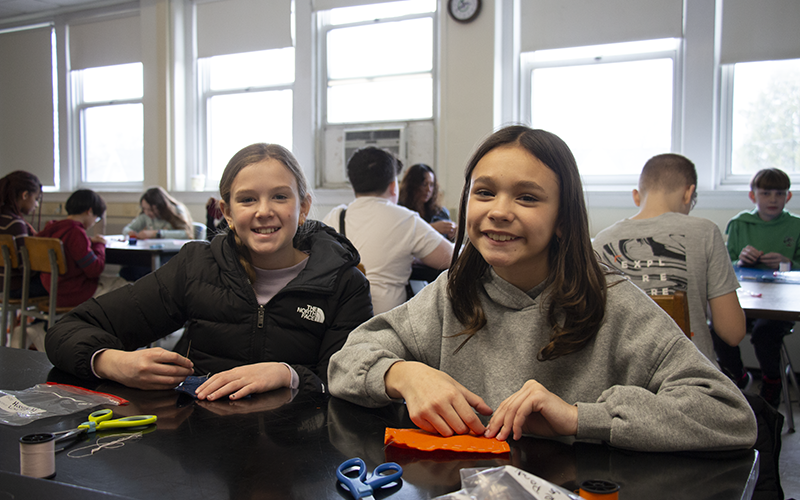 Two middle school girls smile as they sit at a table and sew.