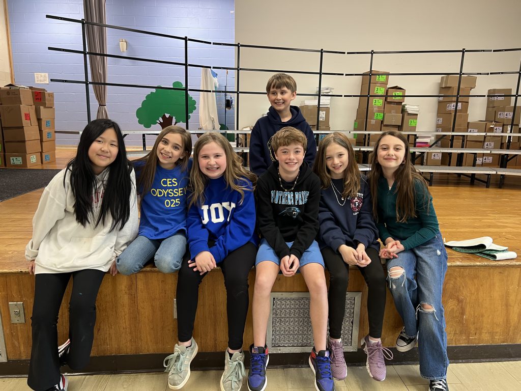 Seven elementary students sit on a stage and smile.