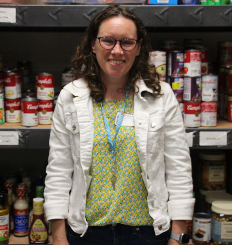 A woman wearing a white jacket and green shirt smiles. Behind her are shelves of canned food.