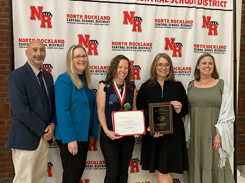 Five people stand infront of a white and red backdrop that says North Rockland. A man on the left and five women. The two women in the center are holding awards.
