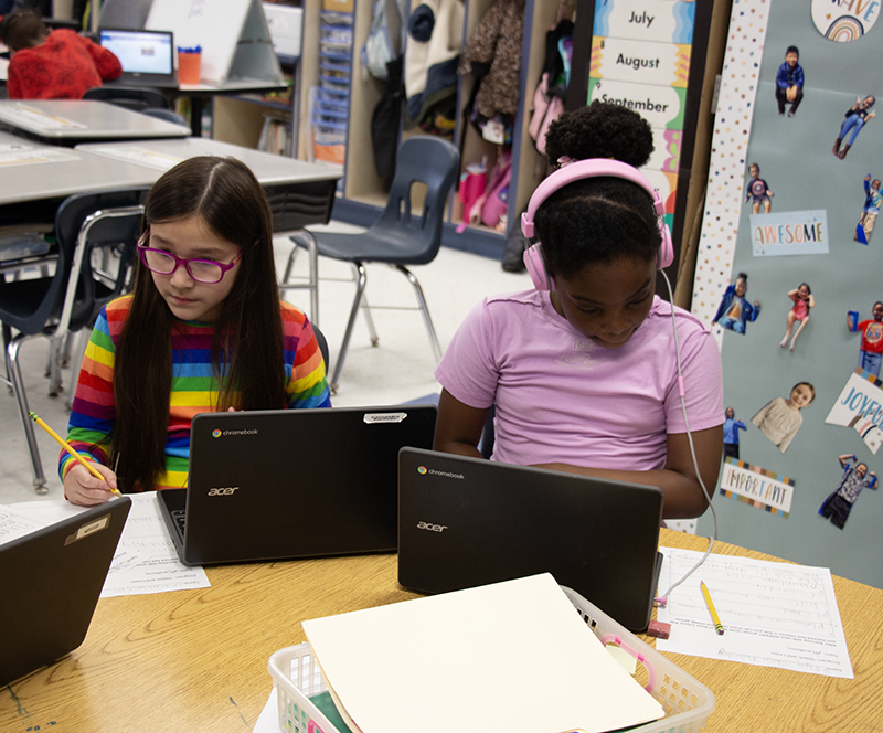 Two second grade girls sit and work on their Chromebooks.