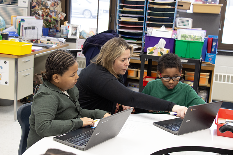 A woman sits in between two second-grade students who are working on Chromebooks. She is pointing to the screen of one of their Chromebooks.