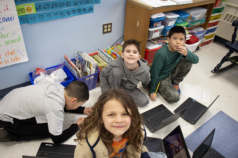 a group of four second-grade kids sit on the floor but look up at the camera. They have Chromebooks they are working on.