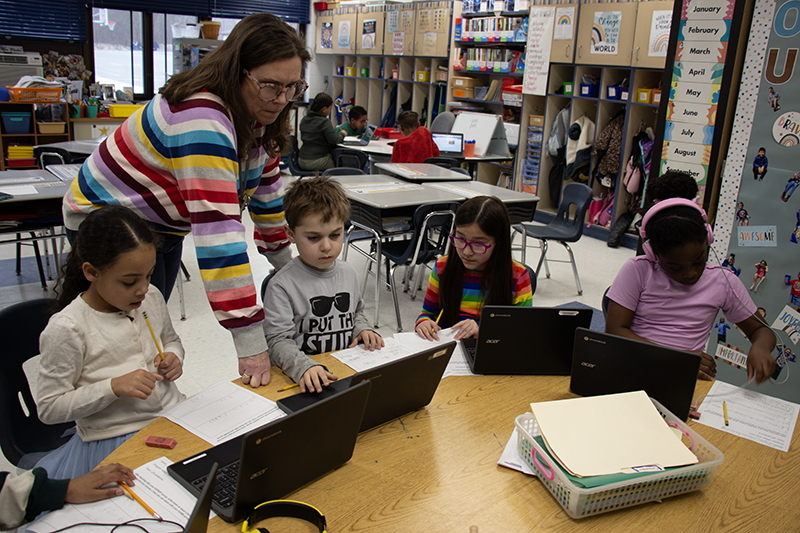 A woman in a  multicolored striped sweater leans in to help second-grade students work on Chromebooks.