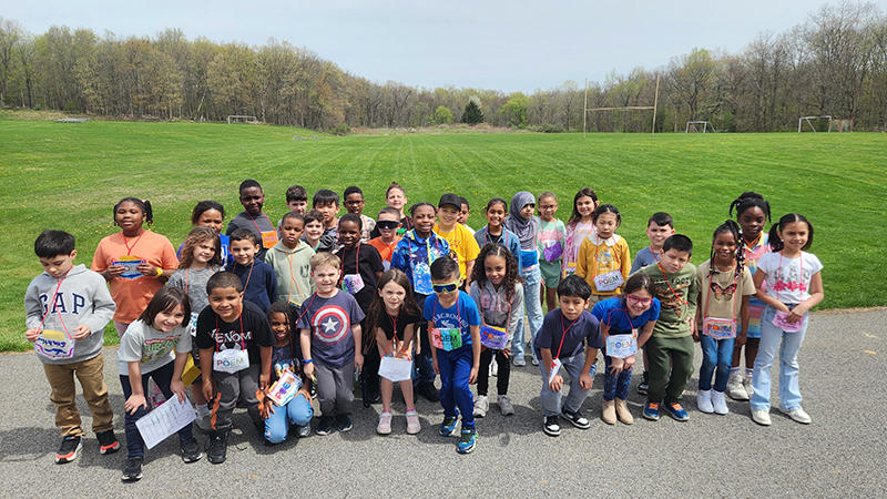 A group of about 36 elementary studnets standin goutside on a bright sunny day, all wearing little pockets on  strings around their necks. The pockets say Poem in My Pocket.