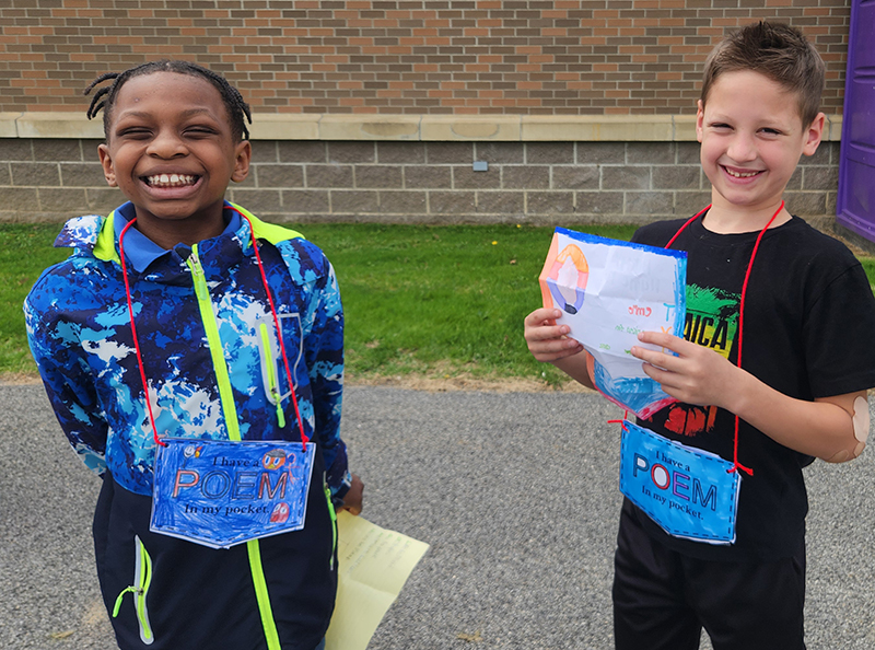 Two elementary age boys smile and hold their poems they are reading to each other.