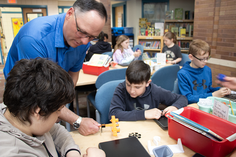 A man stands over a table where three boys are sitting working. He is helping them.