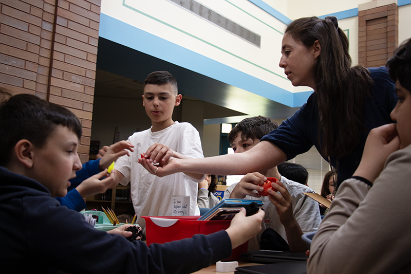 A woman with long dark hair holds out her hand to show kids at a table something.