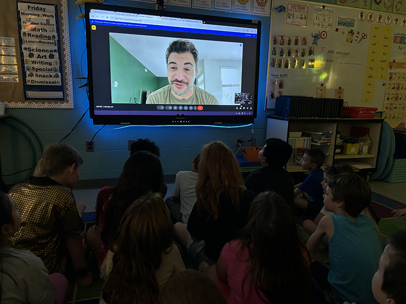 A large screen with a man talking to a large group of elementary students.