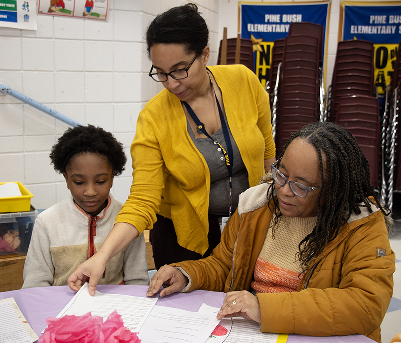 A fifth'grade student sits with a woman and a another woman leans it to work with them.