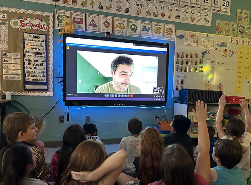 A large screen with a man talking to a group of elementary kids sitting watching. Some kids have their hands up.