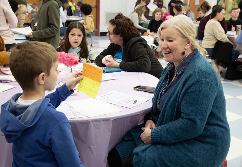 A fifth-grade boy reds a poem to a woman who is sitting and smiling.
