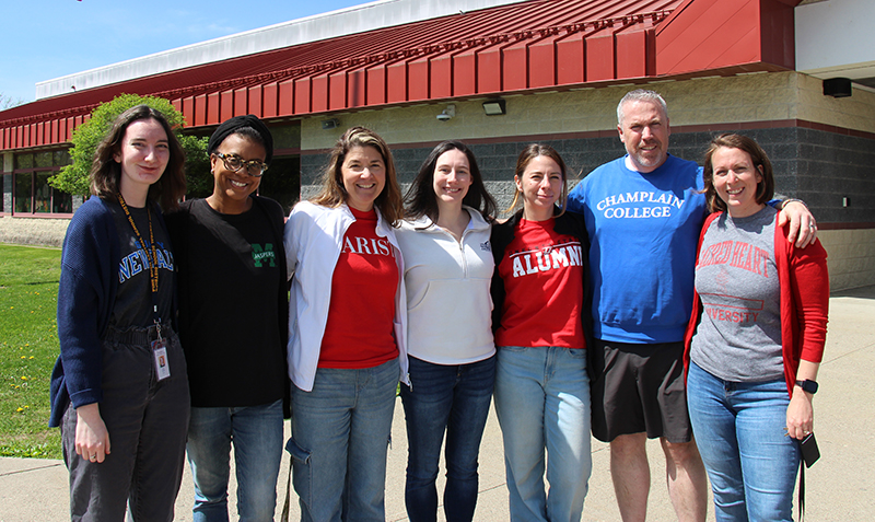 On a sunny day, seven adults stand outside wearing their college tshirts.