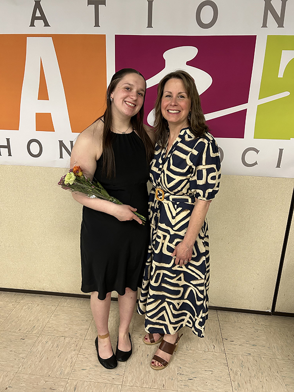 A woman in a black and white print dress stands and smiles with a young woman wearing a black dress and holding a flower.