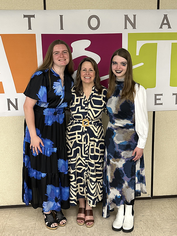 A woman in a black and white print dress stands in between two high school girls who are smiling. They are dressed nicely in dresses.