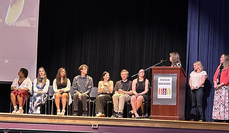 A group of 7 high school students sit on a stage as three women stand to the right. One of the women is speaking behind a podium.