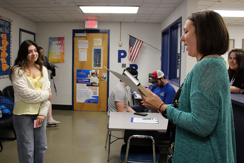 A woman in a green sweater holds a high school yearbook as a young woman listens to her speaking.