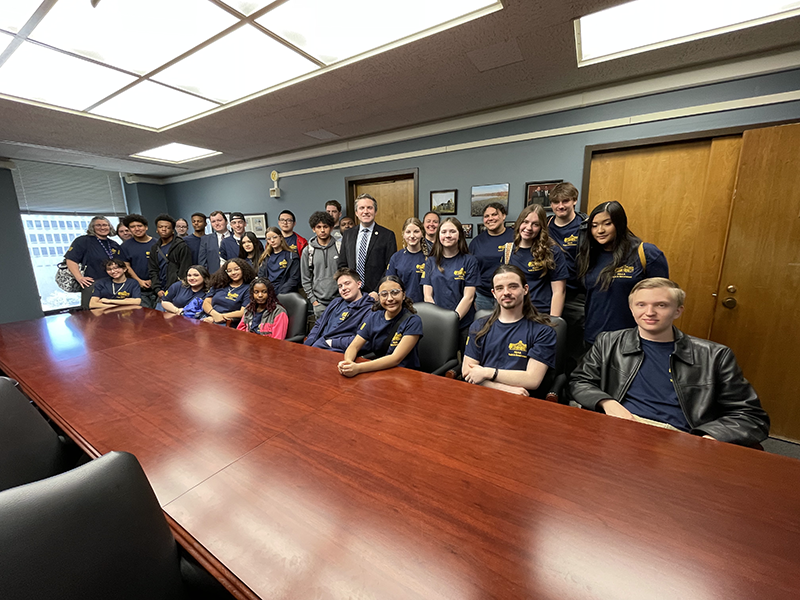 A large group of high school kids sit at a very large conference table with an adult.