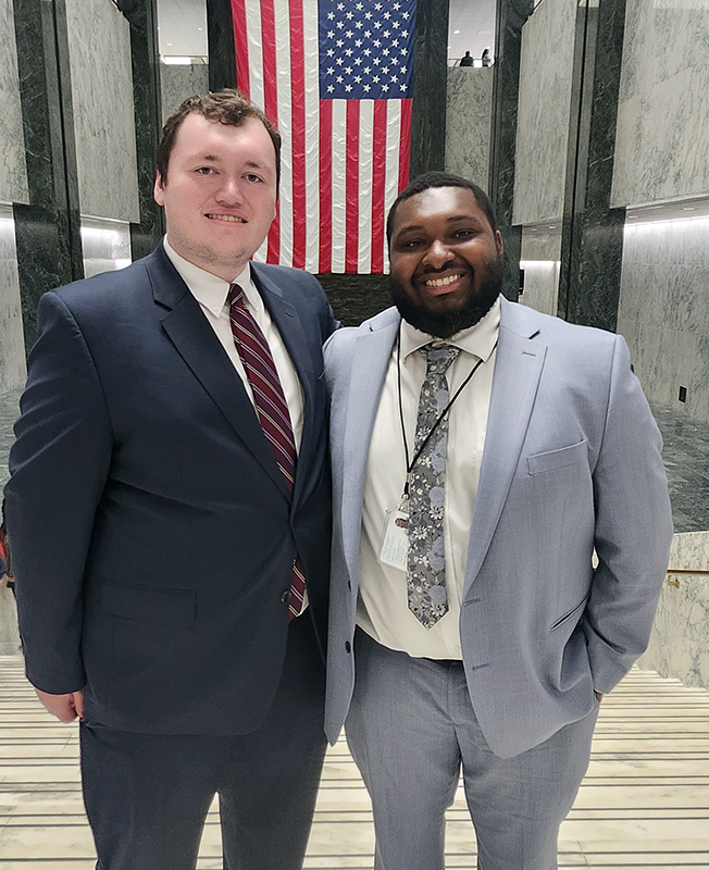 Two young men wearing suits stand together and smile. Behind them is a large American flag.