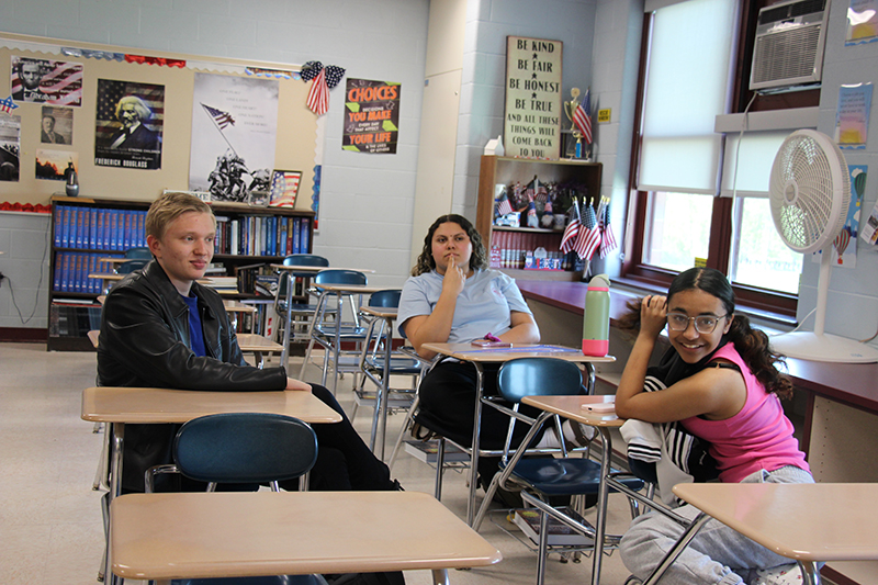Three high school students sit at their desks talking.