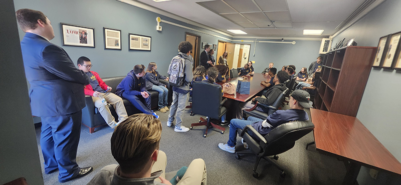 About 25 high school students sit in an office and listen to a man speak.
