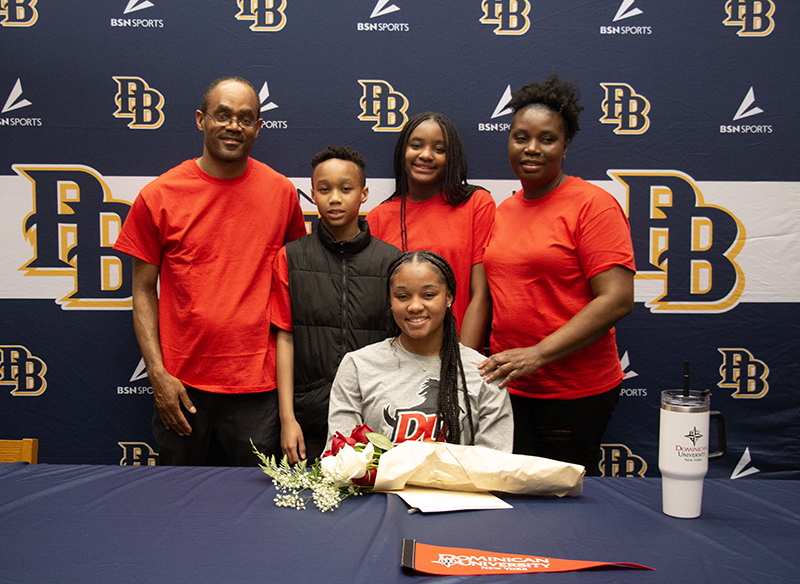 A young woman sits at a table with four people behind her. They are all smiling. There are flowers on the table.