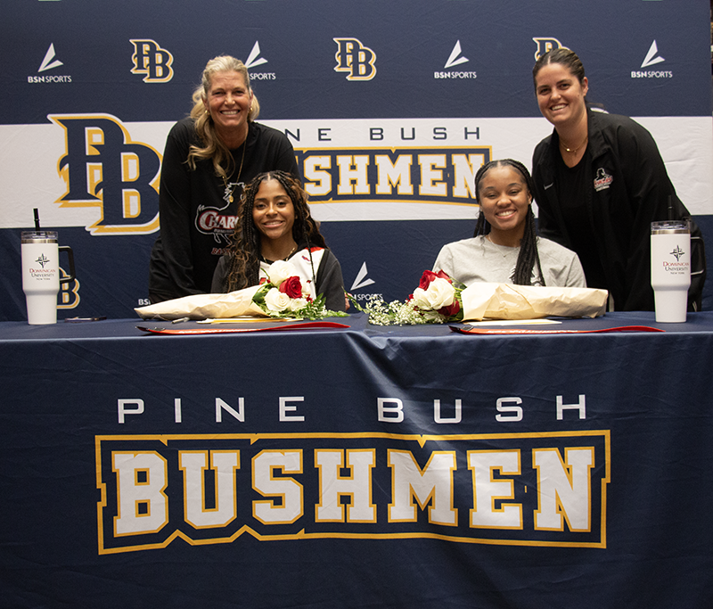 Two young women sit at a table that has a dark blue tablecloth on it that says Pine Bush Bushmen. Behind them are two women smiling.