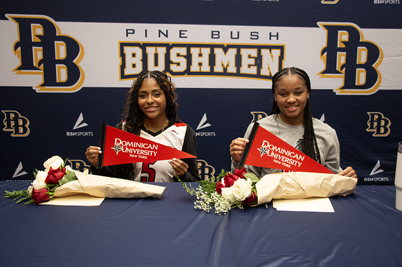 Two young women sit at a table smiling. they are each holding small red pennants that say Dominical University. Behind them a sign says Pine Bush Bushmen.