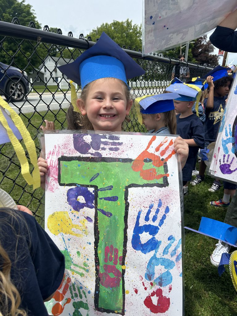 A little girl with a blue graduation cap on holds a sign that has hand prints on it.