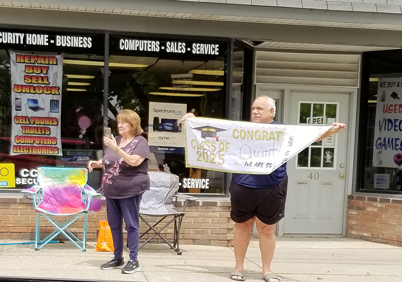 Two adults standing on a sidewalk hold up a sign congratulating the graduates.