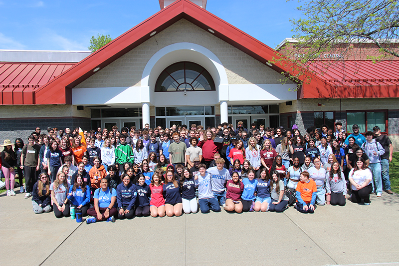 A very large group of high school seniors stand and squat outside their school building , most wearing their college t shirts.